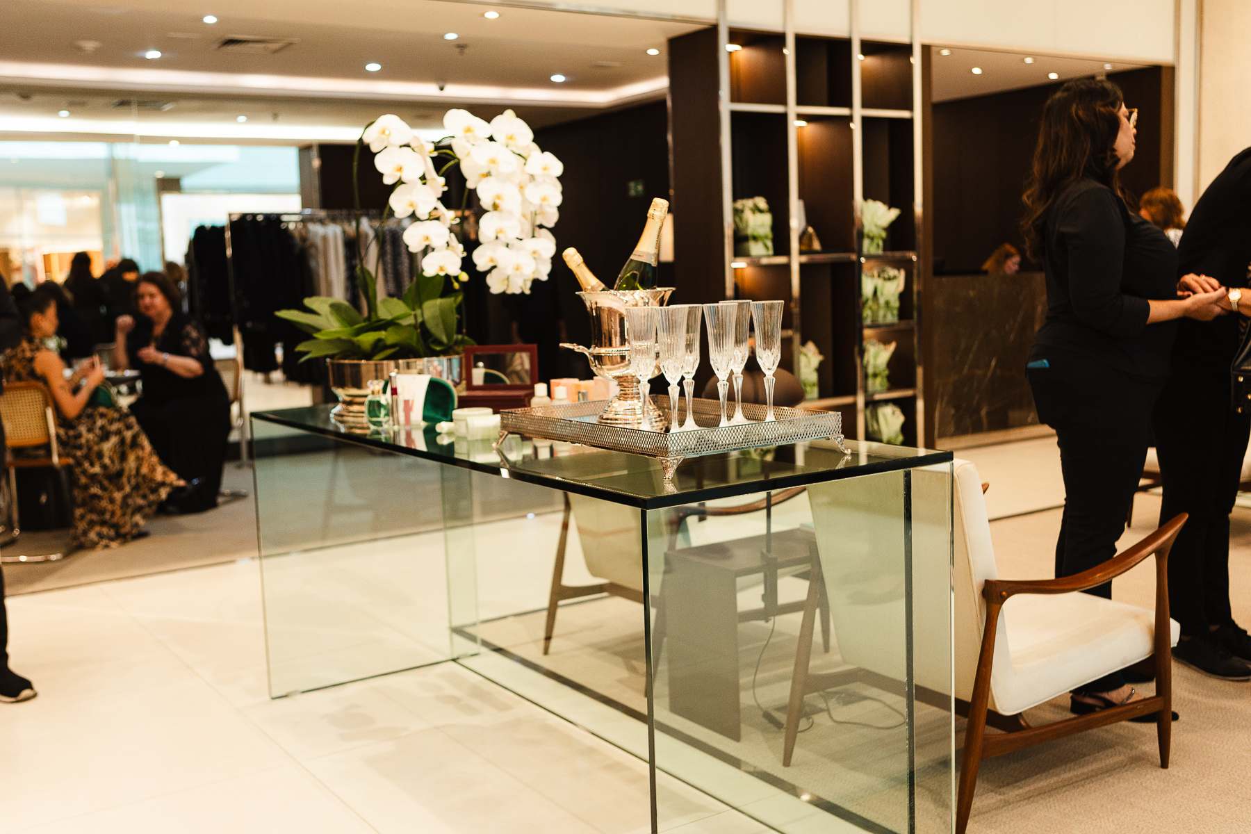 Recepção VIP com balde de prata com champanhe no gelo, taças sobre bandeja prateada, orquídeas brancas e cosméticos em uma mesa de vidro, com convidados ao fundo
