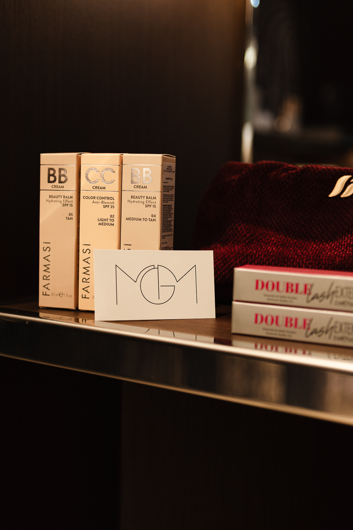 Caixas de BB e CC Cream Farmasi, cartão com o monograma MGM, nécessaire bordô e máscaras Double Lash dispostos sobre uma prateleira escura com filete cromado