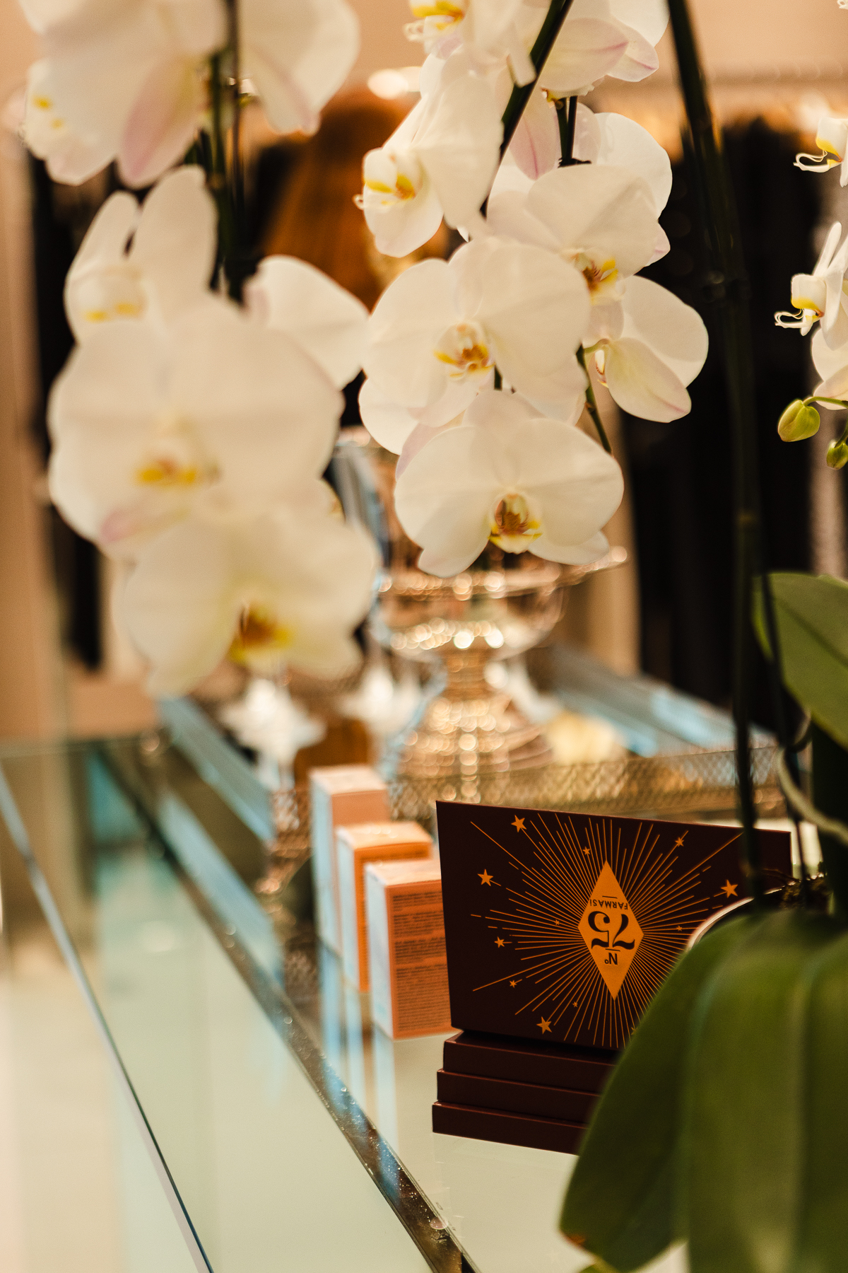 Vitrine de produtos Farmasi com orquídeas brancas e embalagem bordô N° 75 sobre bancada de vidro