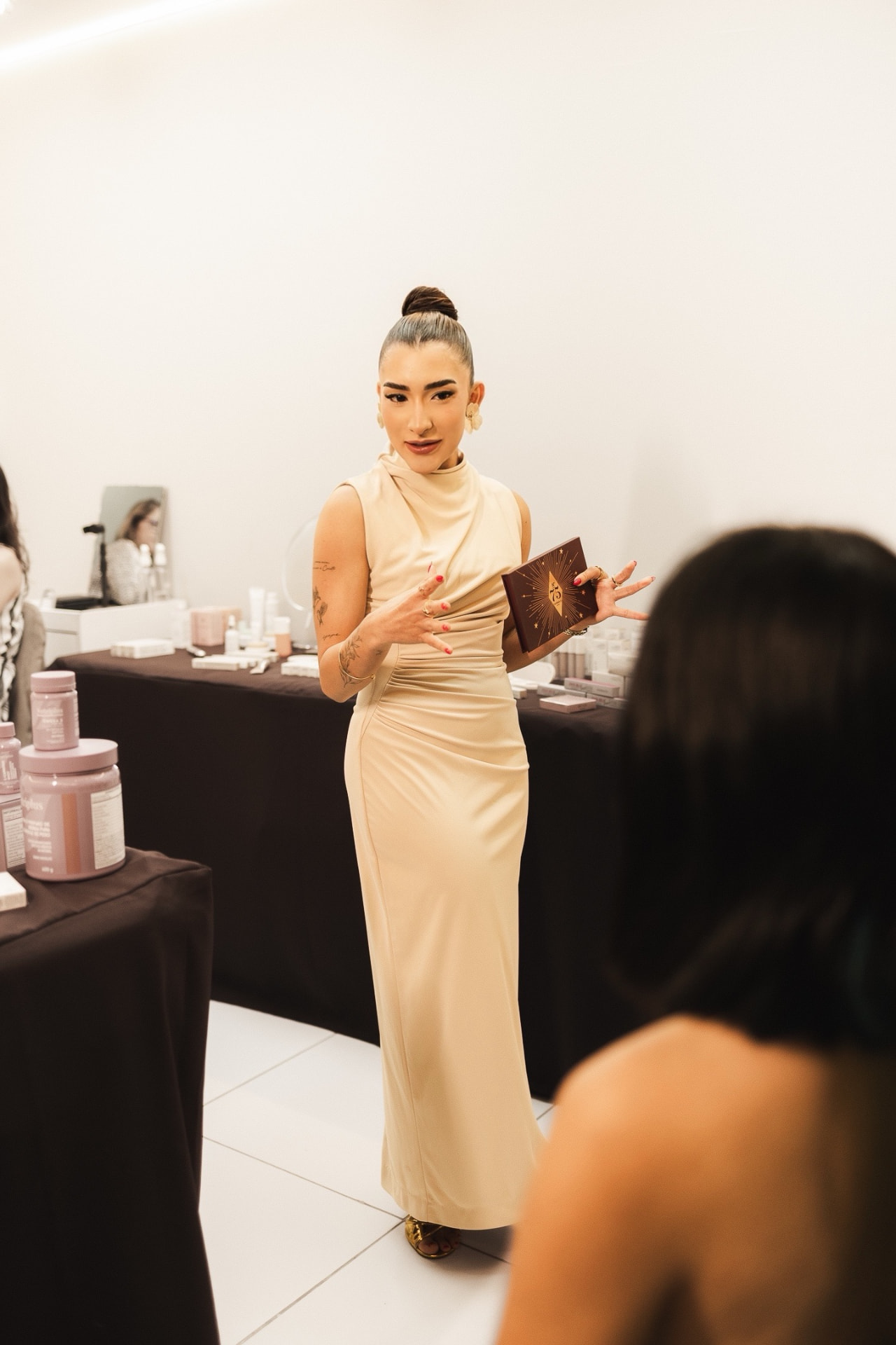 Maria Gabriela, de vestido claro, apresentando uma paleta de maquiagem a uma cliente sentada à sua frente, em uma estação de maquiagem da MGM