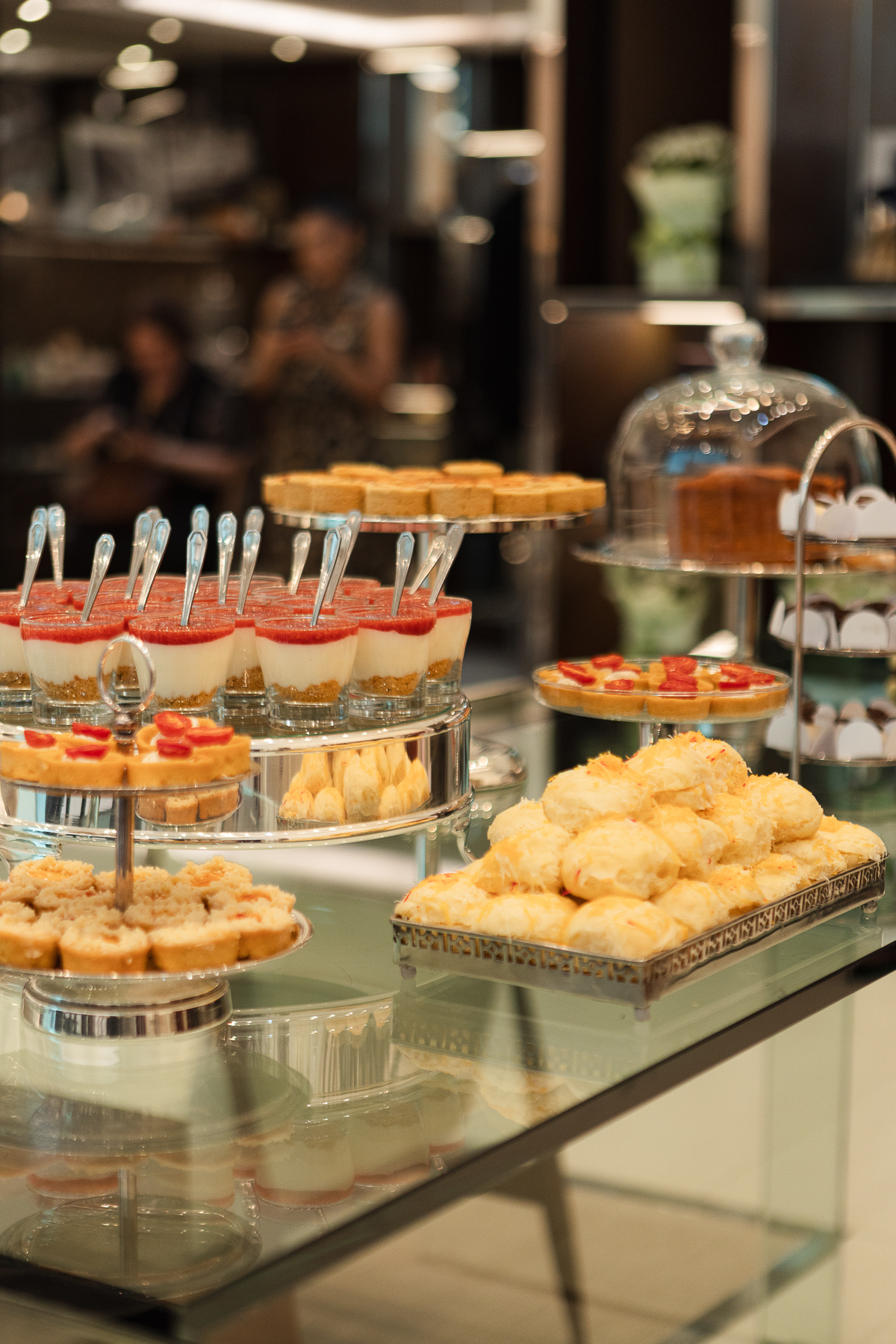 Mesa de buffet com copinhos de sobremesa, mini tortinhas em suportes de prata e bandeja de salgados, com convidados ao fundo