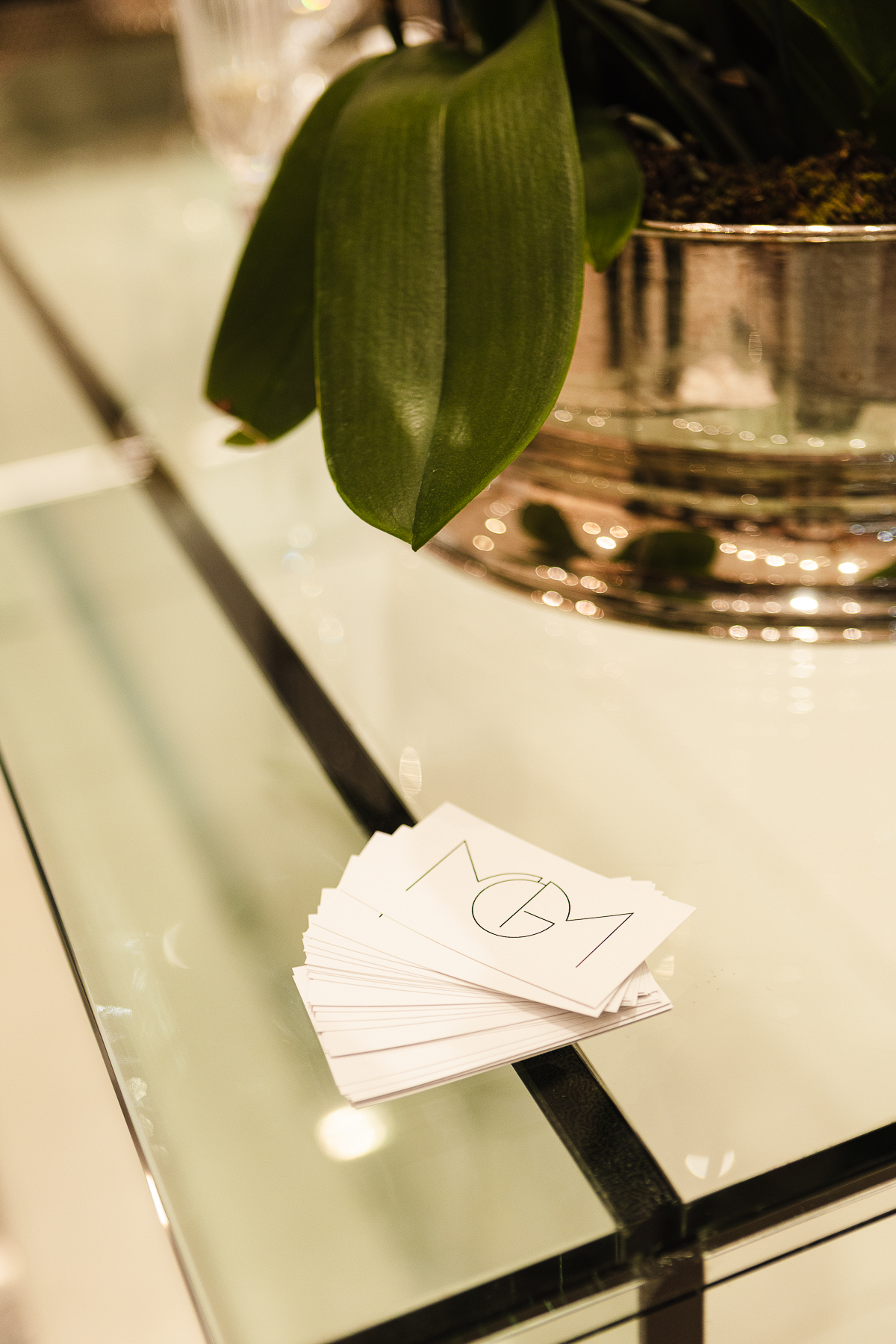 Cartões de visita MGM com monograma, sobre tampo de vidro, ao lado de folhas de orquídea em vaso de metal