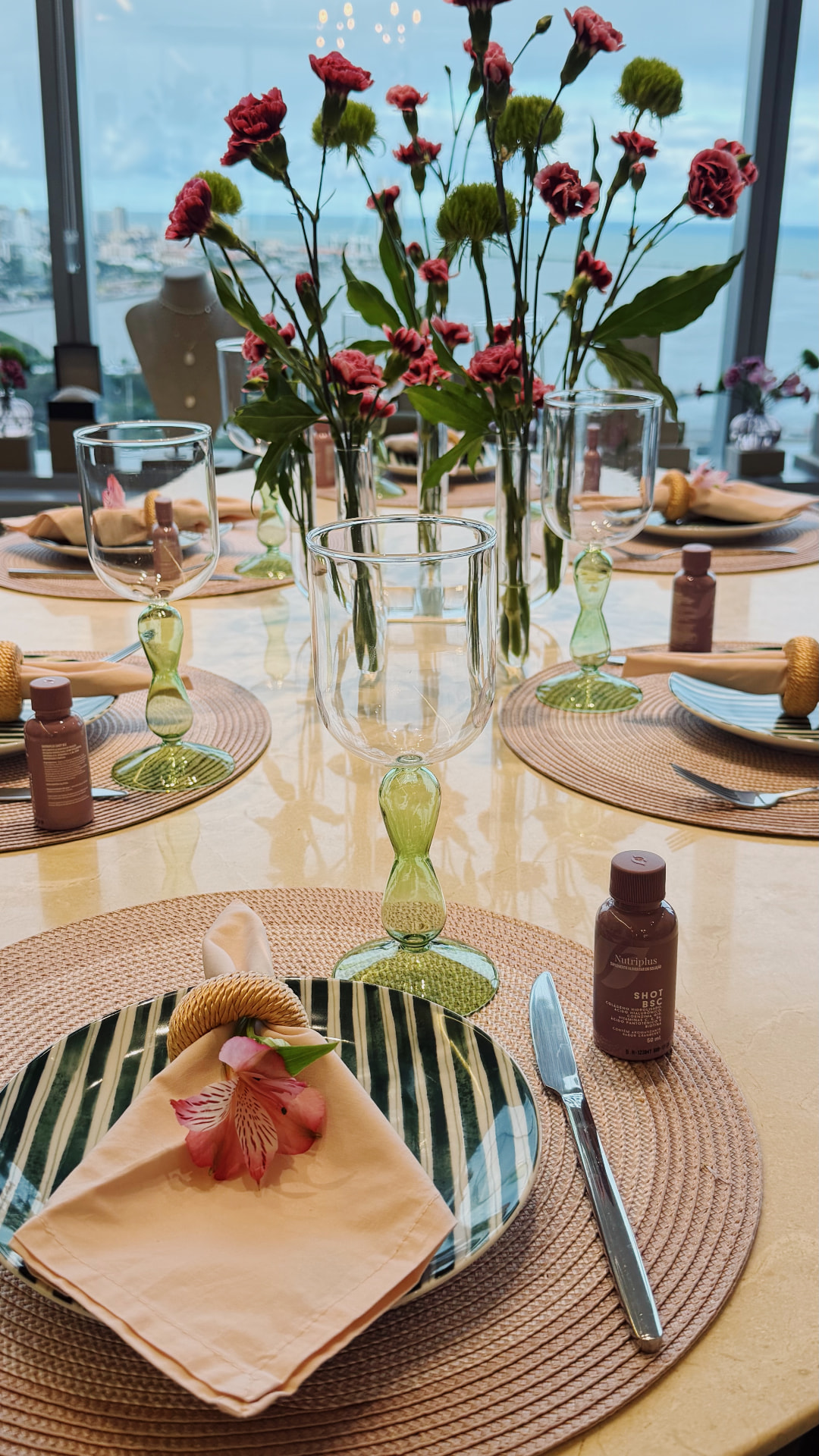 Mesa posta para evento com sousplats de fibra, guardanapos salmão, taças de haste verde e arranjo floral alto