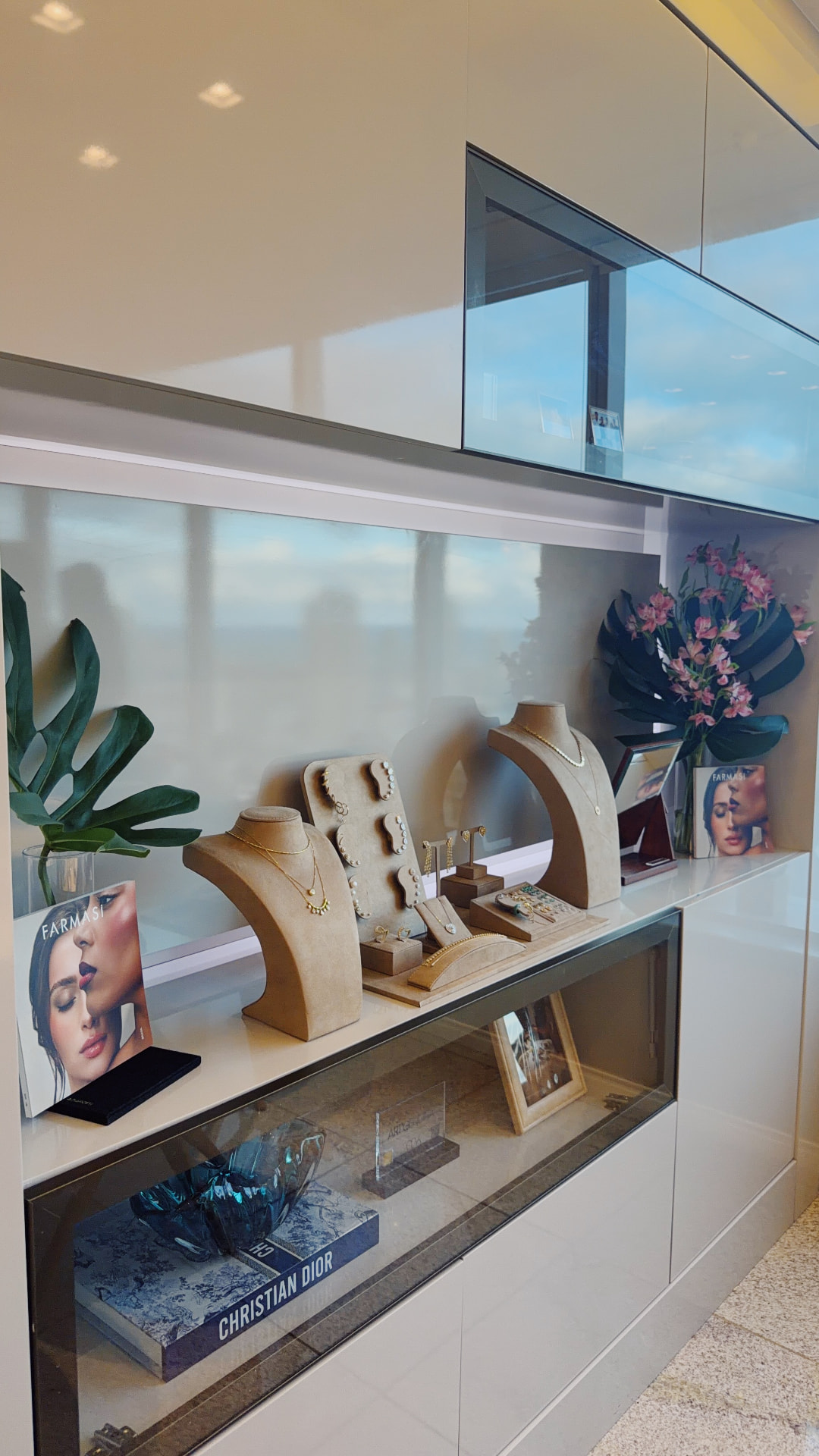 Vitrine da Maison com bustos de joias em camurça bege, folhetos Farmasi, arranjos florais e um livro de arte na vitrine de vidro